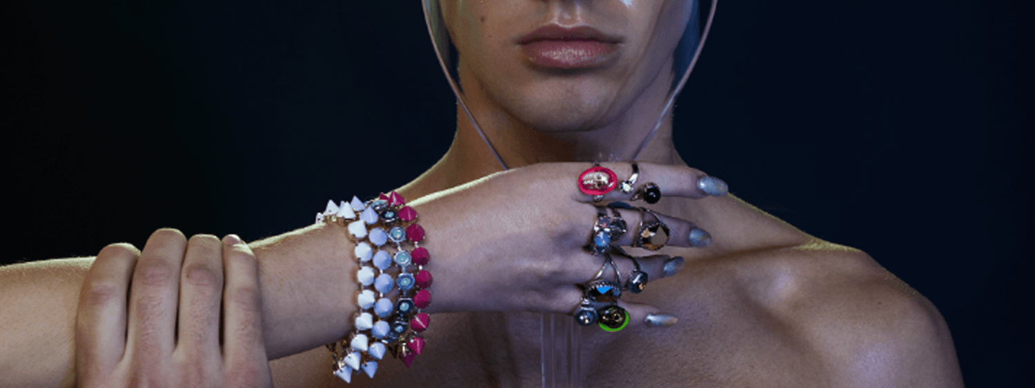 A hand decked out with spiked bracelets, crystal rings, skull jewelry holding a crystal clear faceshield in front of a male model.