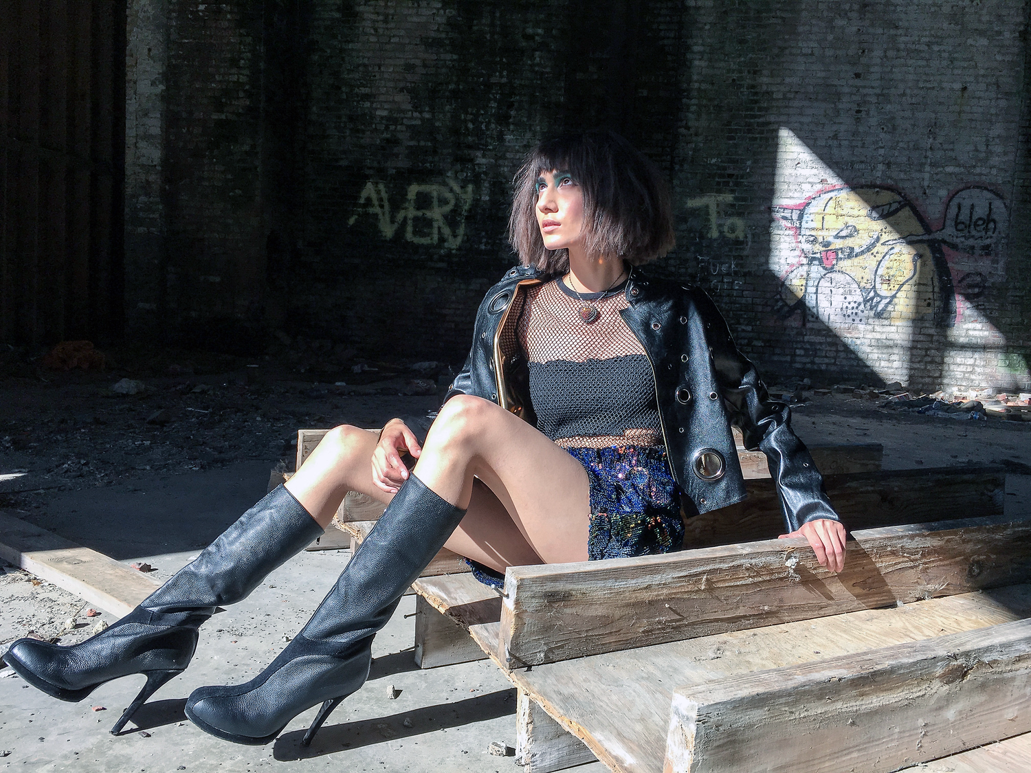 Model wearing a black tube top with a fishnet shirt underneath a leather jacket and thigh high boots. She is looking up towards the sun which catches the sparkle of the Bunny Paige Spiked and Paved Heart she's wearing around her neck.